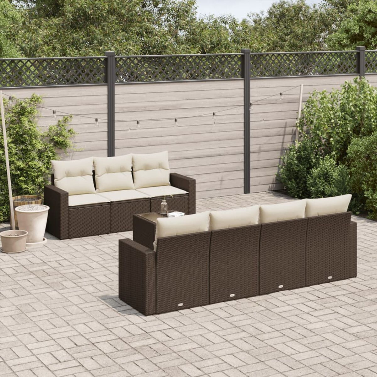 Set divani da esterno in vimini marrone con cuscini beige, su un patio. Piante verdi e recinzione sullo sfondo.