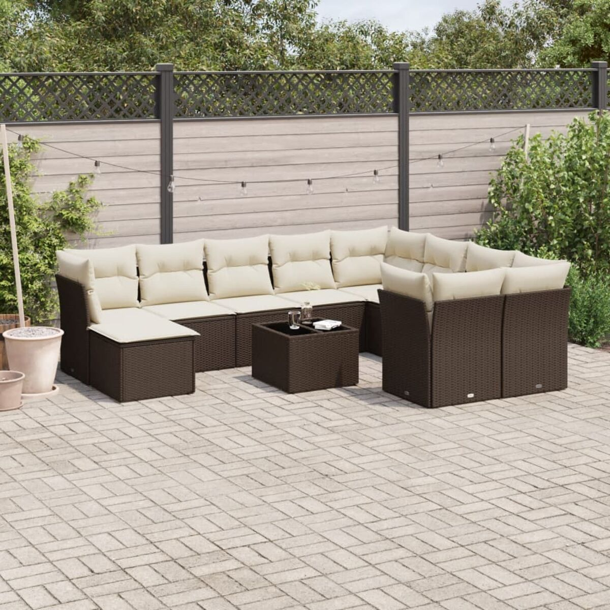 Divano angolare in vimini marrone con cuscini color crema, tavolino, su patio. Verde e recinzione sullo sfondo.