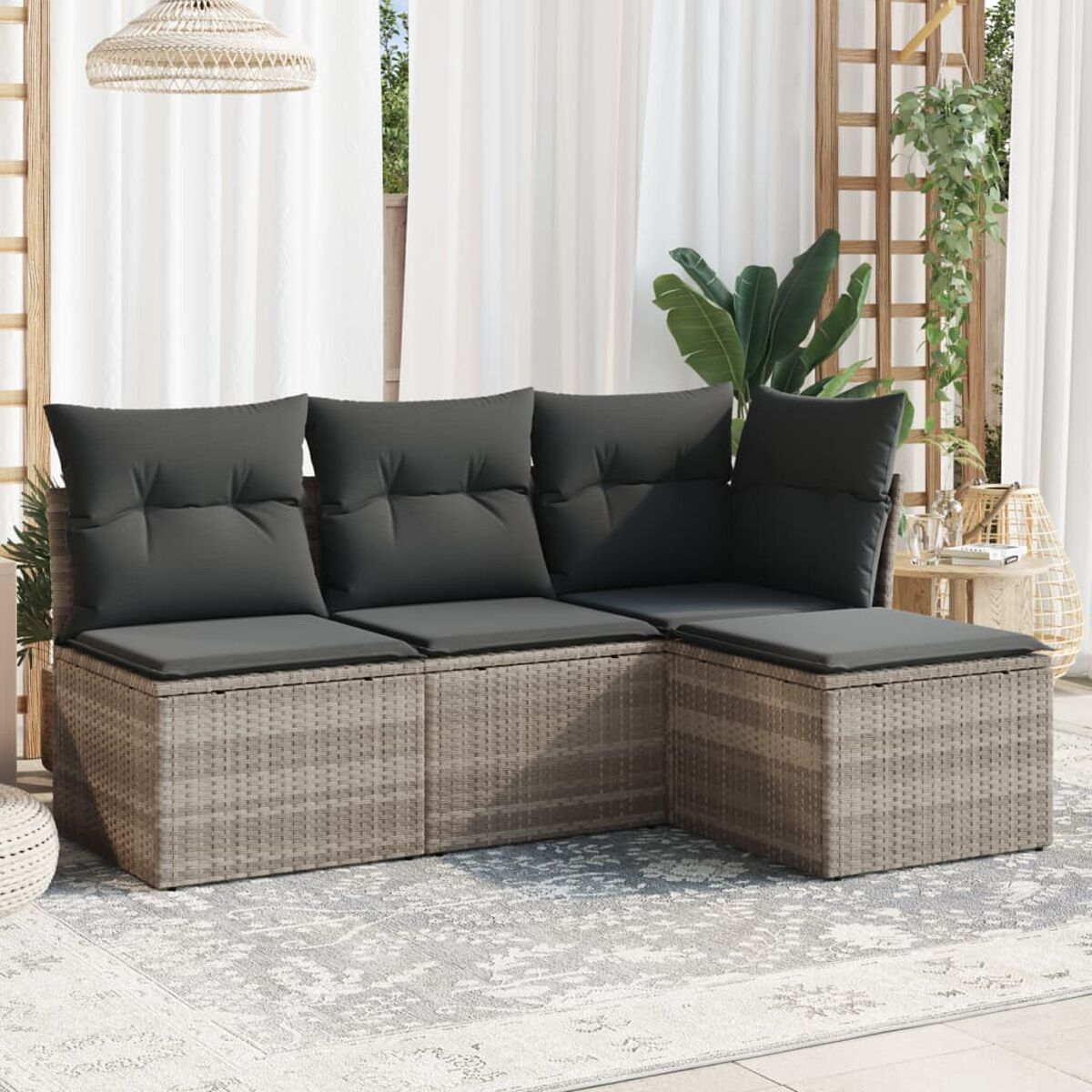 Divano a L in rattan con cuscini grigi, su un tappeto fantasia. Tende e piante visibili sullo sfondo.