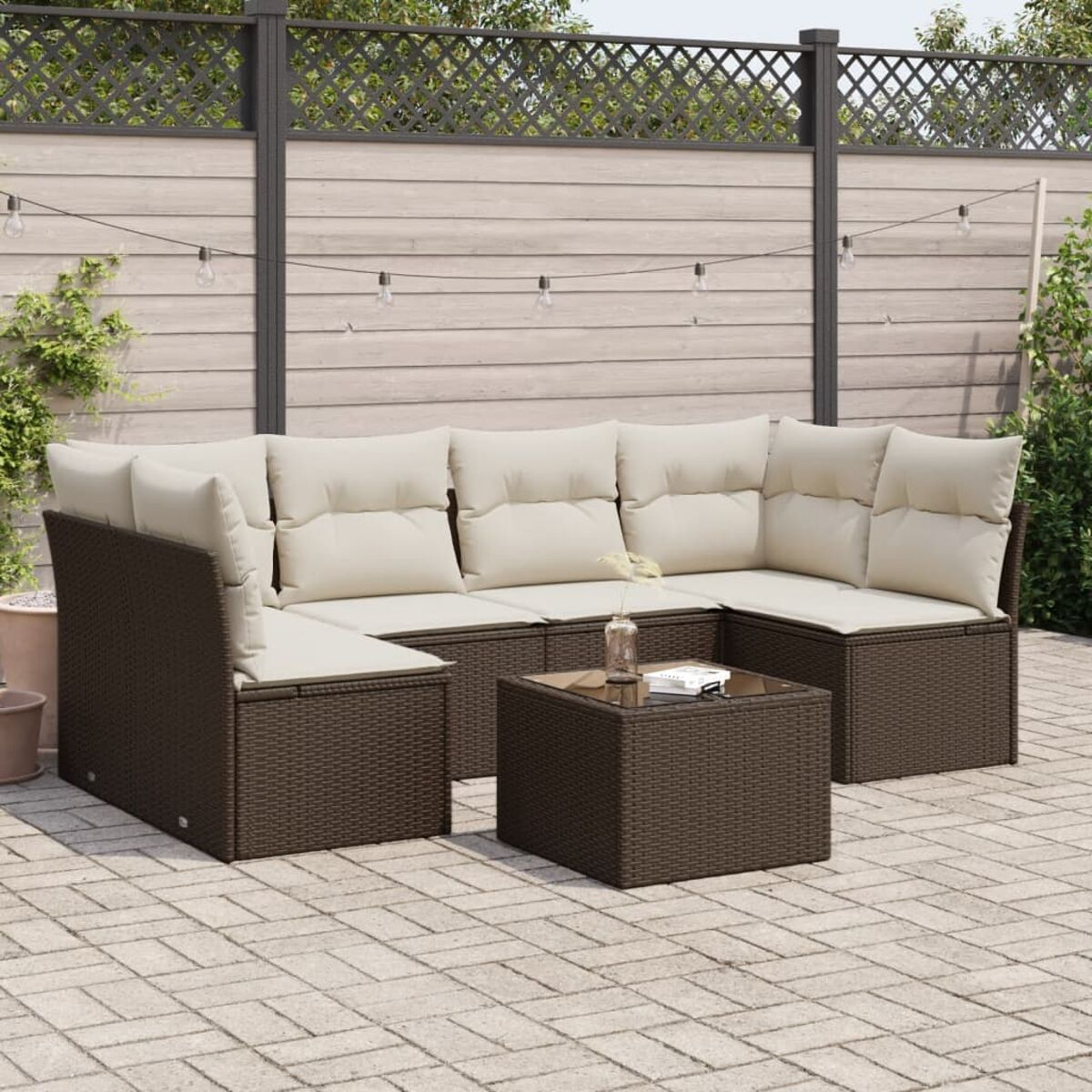 Set da patio in vimini scuro con cuscini color crema, tavolino e luci a corda. Recinzione in legno e pavimentazione.