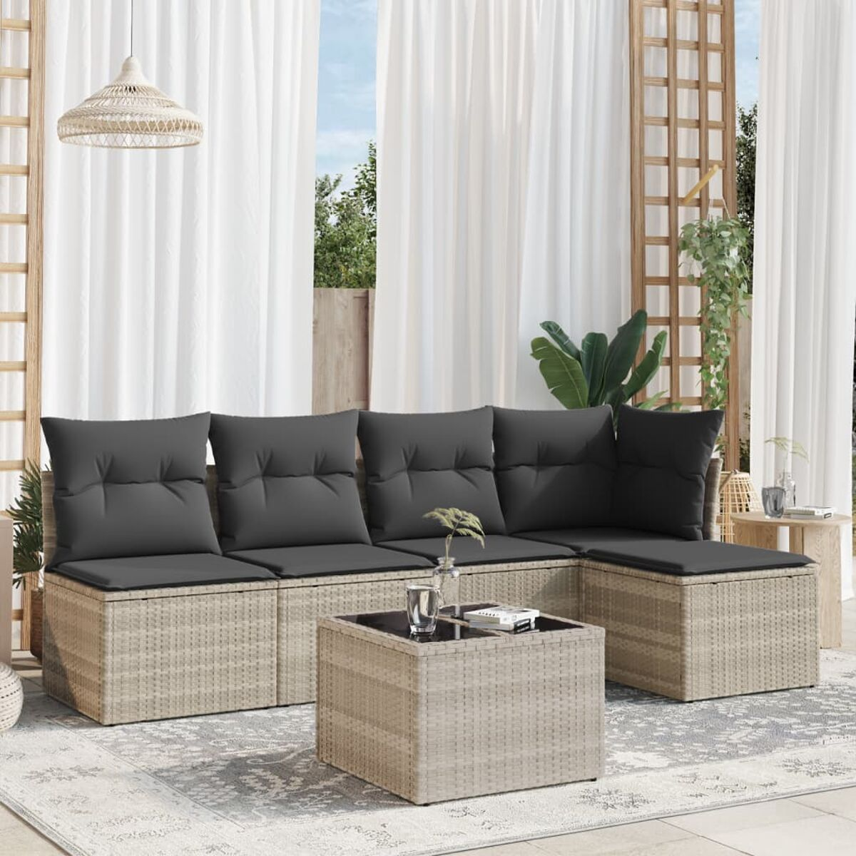 Rattan-Ecksofa mit grauen Kissen, Couchtisch auf einem Teppich. Weiße Vorhänge und Pflanzen im Hintergrund.