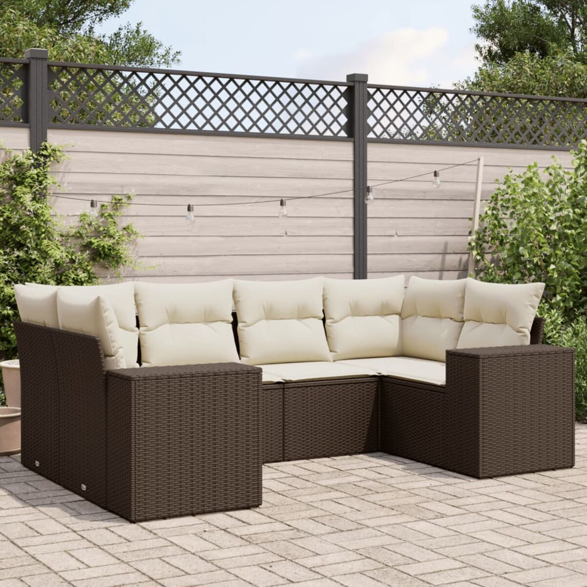 Braunes Korbmöbel-Ecksofa mit cremefarbenen Kissen im Garten. Hintergrund: Zaun, Pflanzen.