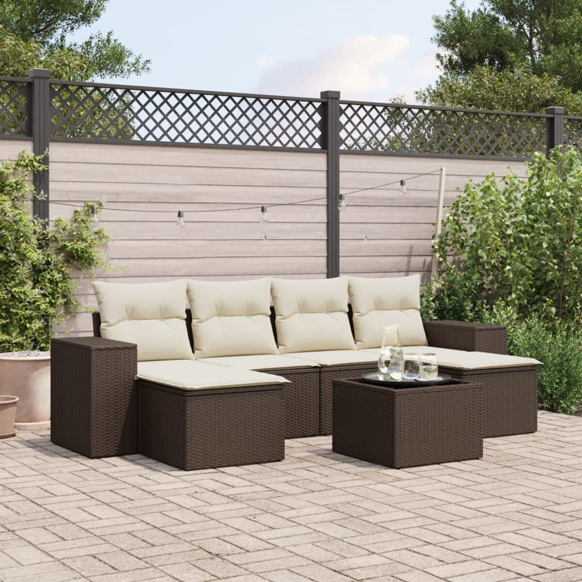 Set divano da esterno in vimini marrone con cuscini beige e tavolino. Patio con pavimentazione in mattoni grigi e una recinzione.