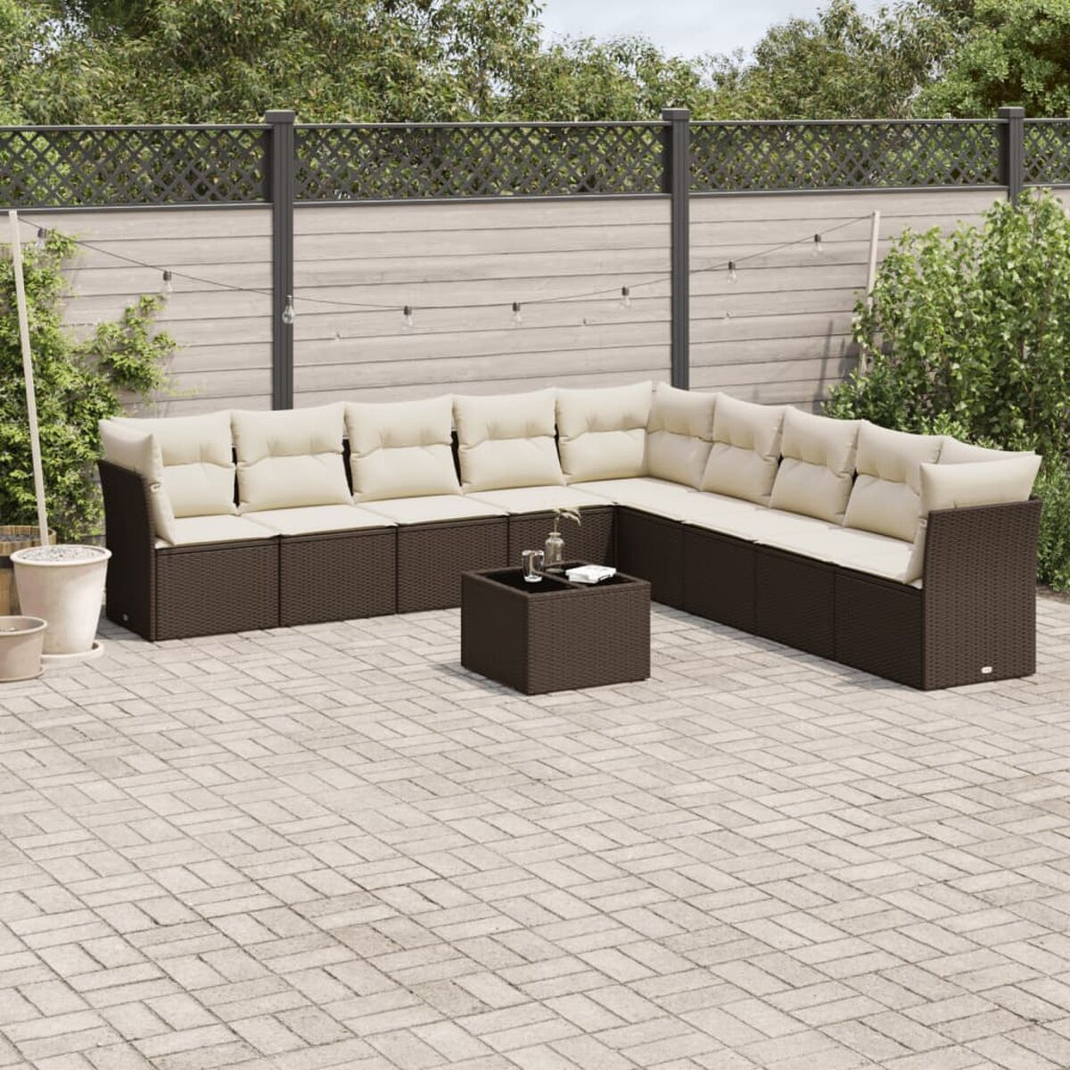 Divano componibile marrone da esterno con cuscini beige su un patio pavimentato. Un tavolo al centro. Sfondo di piante.