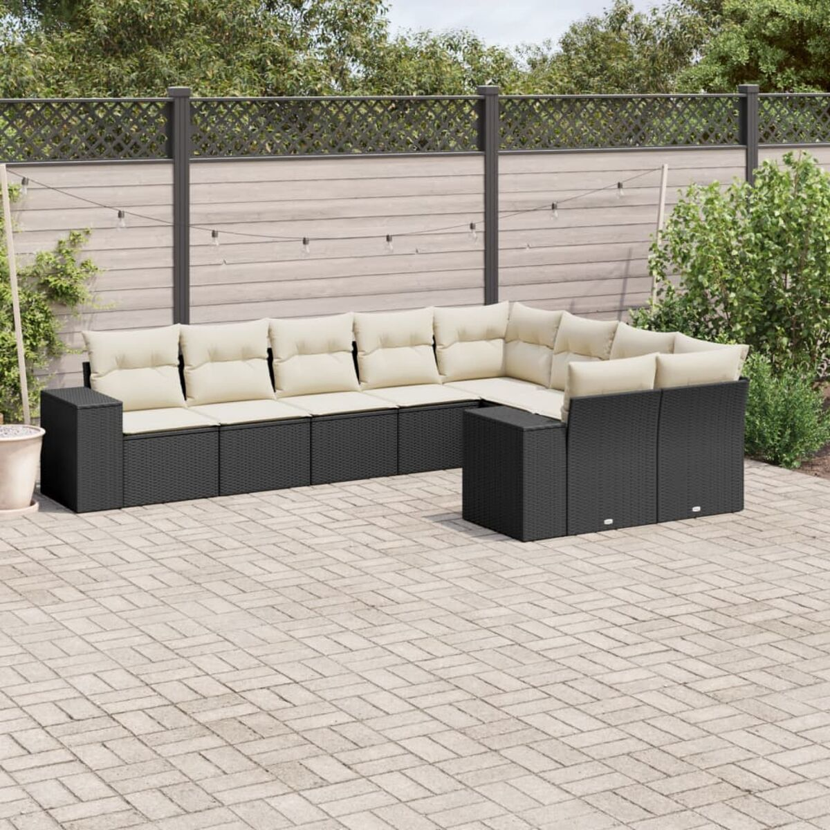 Schwarzes L-förmiges Outdoor-Sofa aus Korbgeflecht mit beigen Kissen auf einer Backstein-Terrasse. Zaun und Pflanzen im Hintergrund.