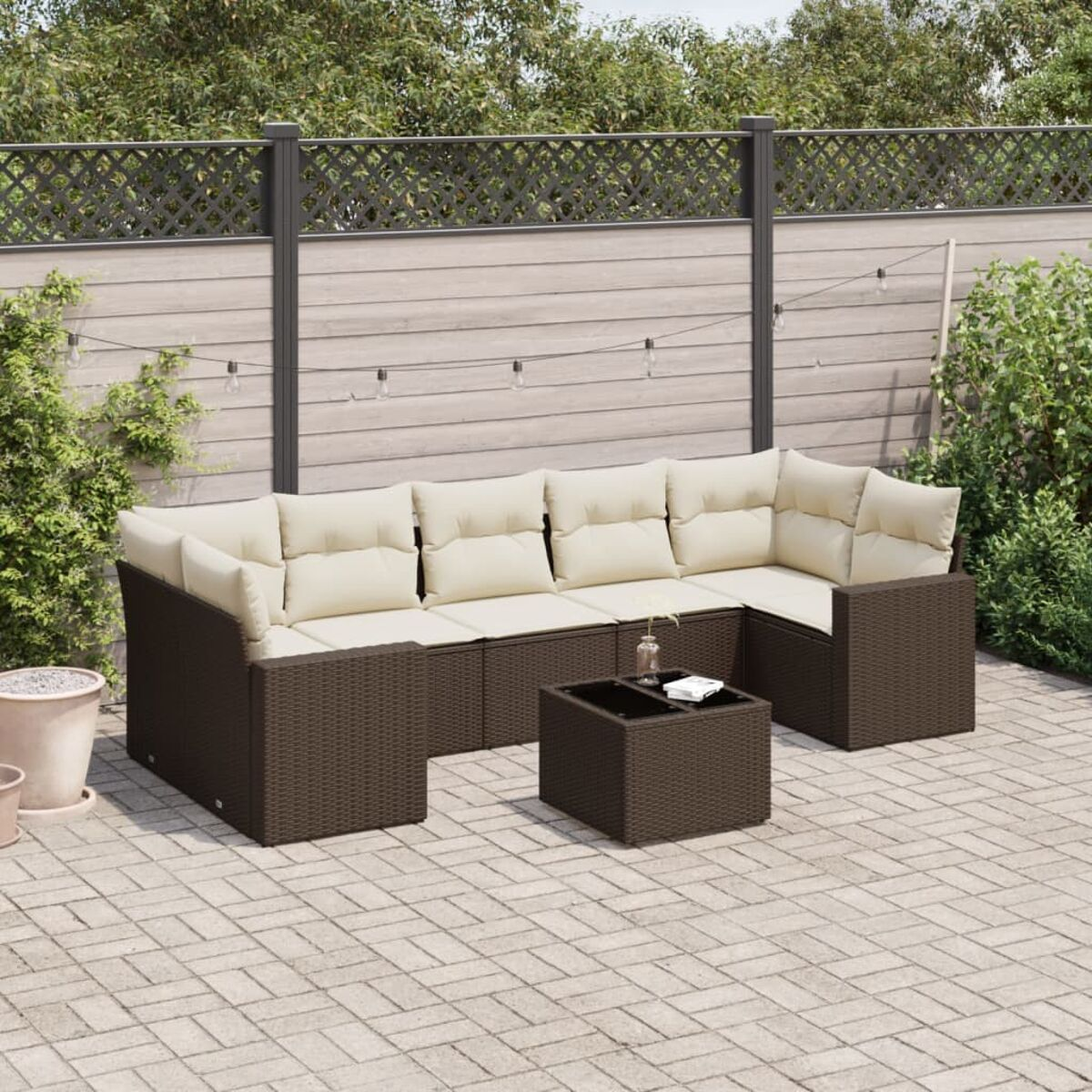 Braunes Ecksofa aus Korbgeflecht mit beigefarbenen Kissen und einem Tisch im Freien. Terrasse mit Zaun und Pflanzen.