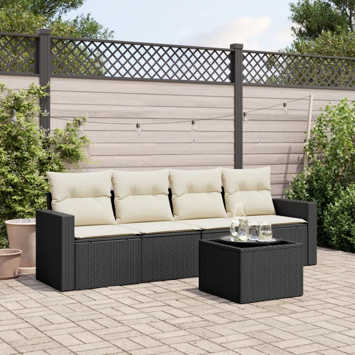 Schwarzes Rattan-Outdoor-Sofa mit cremefarbenen Kissen, Couchtisch mit Getränken auf Ziegelterrasse.