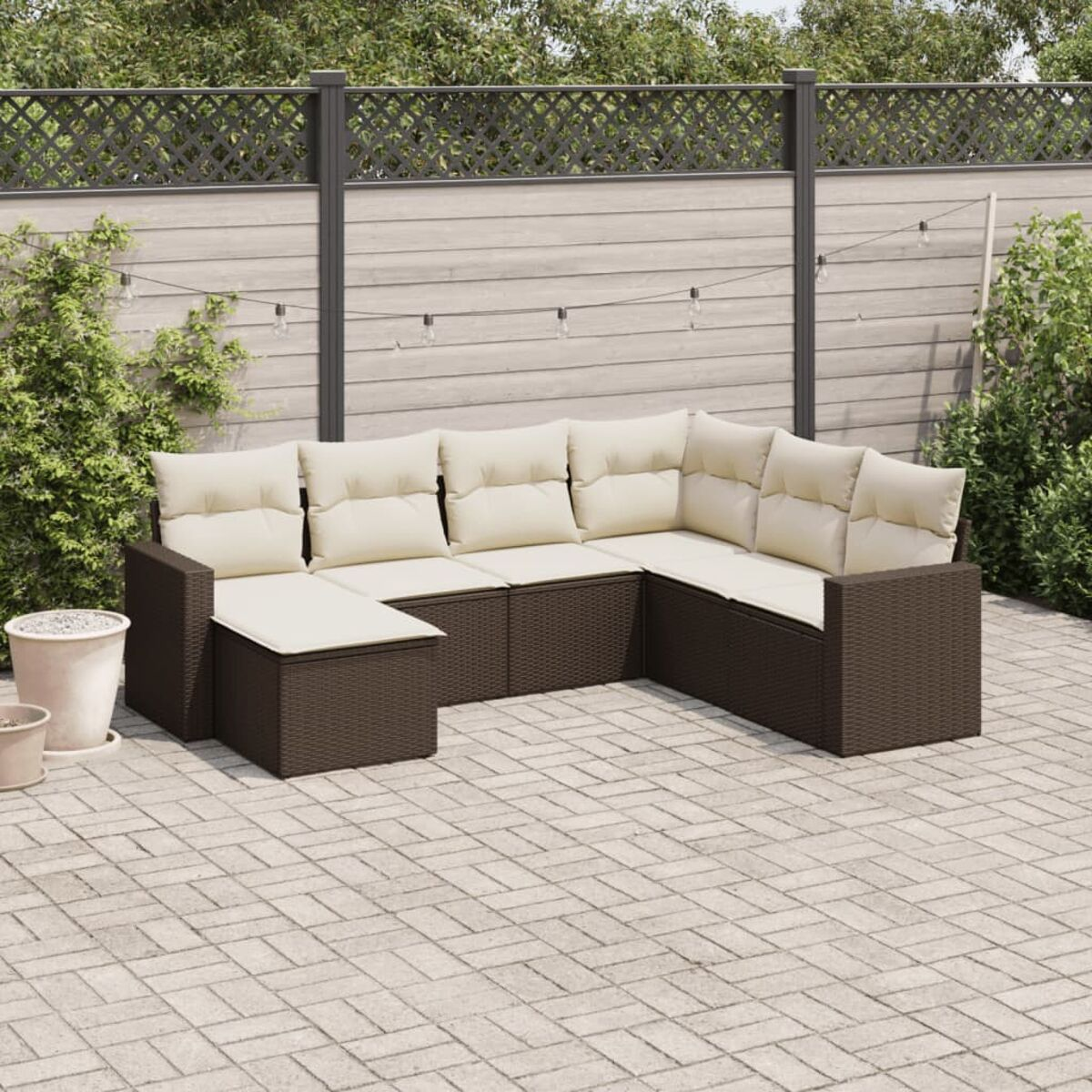Braunes Outdoor-Sofa aus Korbgeflecht mit beigen Kissen. Garteneinstellung mit Pflanzen, Zaun und Lichtern im Hintergrund.