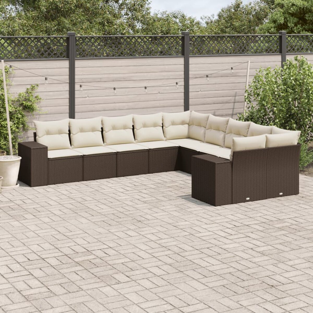 Divano componibile da esterno in vimini marrone con cuscini beige. Su un patio in mattoni con vegetazione.
