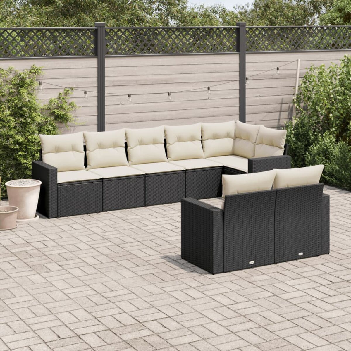 Schwarzes Rattan-Ecksofa im Freien mit beigen Kissen auf gepflasterter Terrasse. Zaun und Pflanzen im Hintergrund.