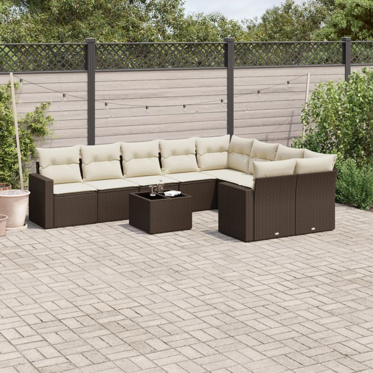 Divano componibile in vimini marrone con cuscini color crema, tavolo quadrato su un patio in mattoni e una recinzione.