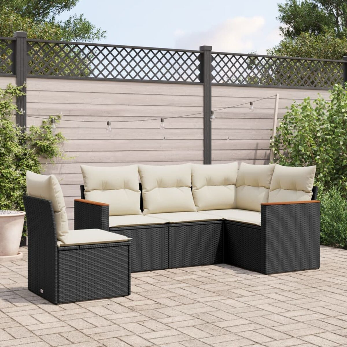 Schwarzes Outdoor-Sofa-Set aus Korbgeflecht mit beigen Kissen und einem Stuhl, auf einer Backstein-Terrasse. Zaun und Pflanzen sichtbar.