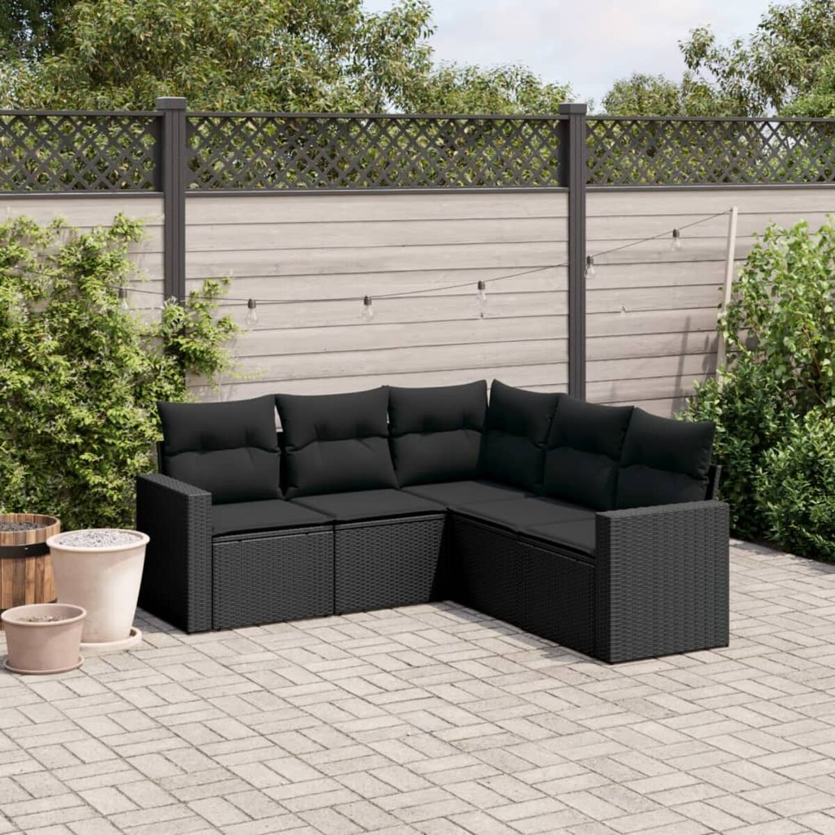 Schwarzes Outdoor-Ecksofa auf einer Backstein-Terrasse, mit Zaun und Pflanzen im Hintergrund.
