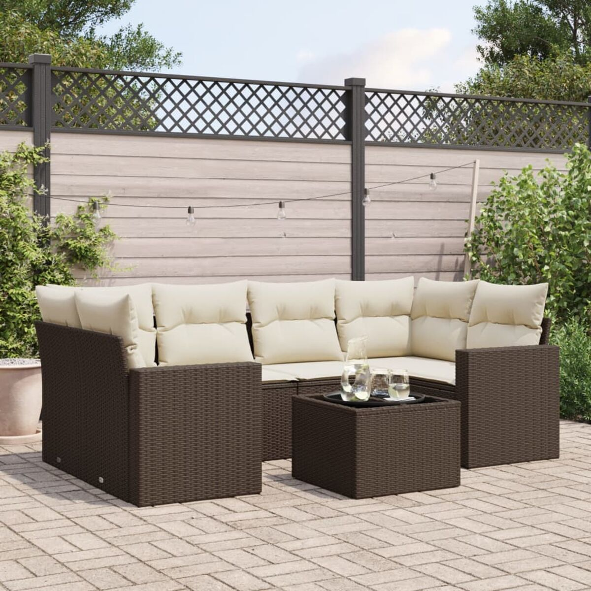 Divano angolare da esterno in vimini marrone scuro con cuscini beige e un tavolino. Patio in mattoni, piante e tende.