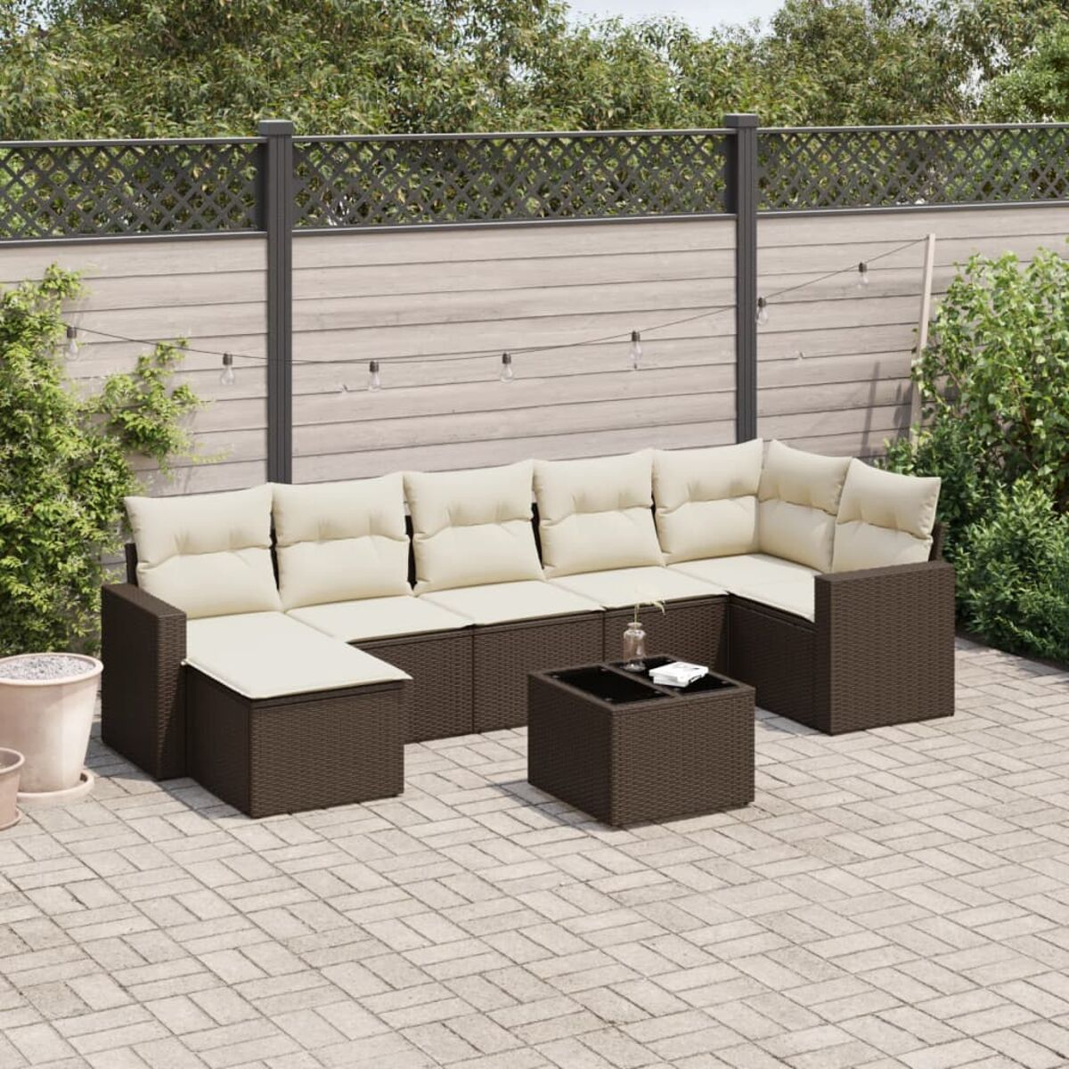 Divano angolare da esterno in vimini marrone con cuscini color crema e un tavolo, su un patio. Sfondo con recinzione e piante.