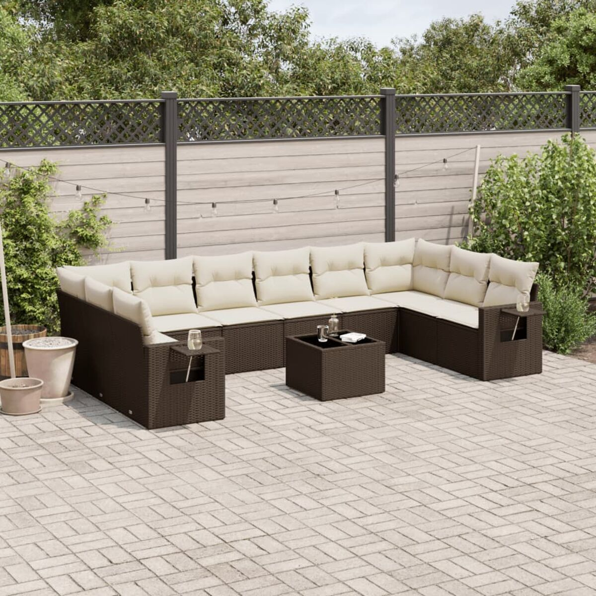 Set divano in vimini marrone con cuscini color crema, tavolo su un patio in mattoni. Verde e recinzione.