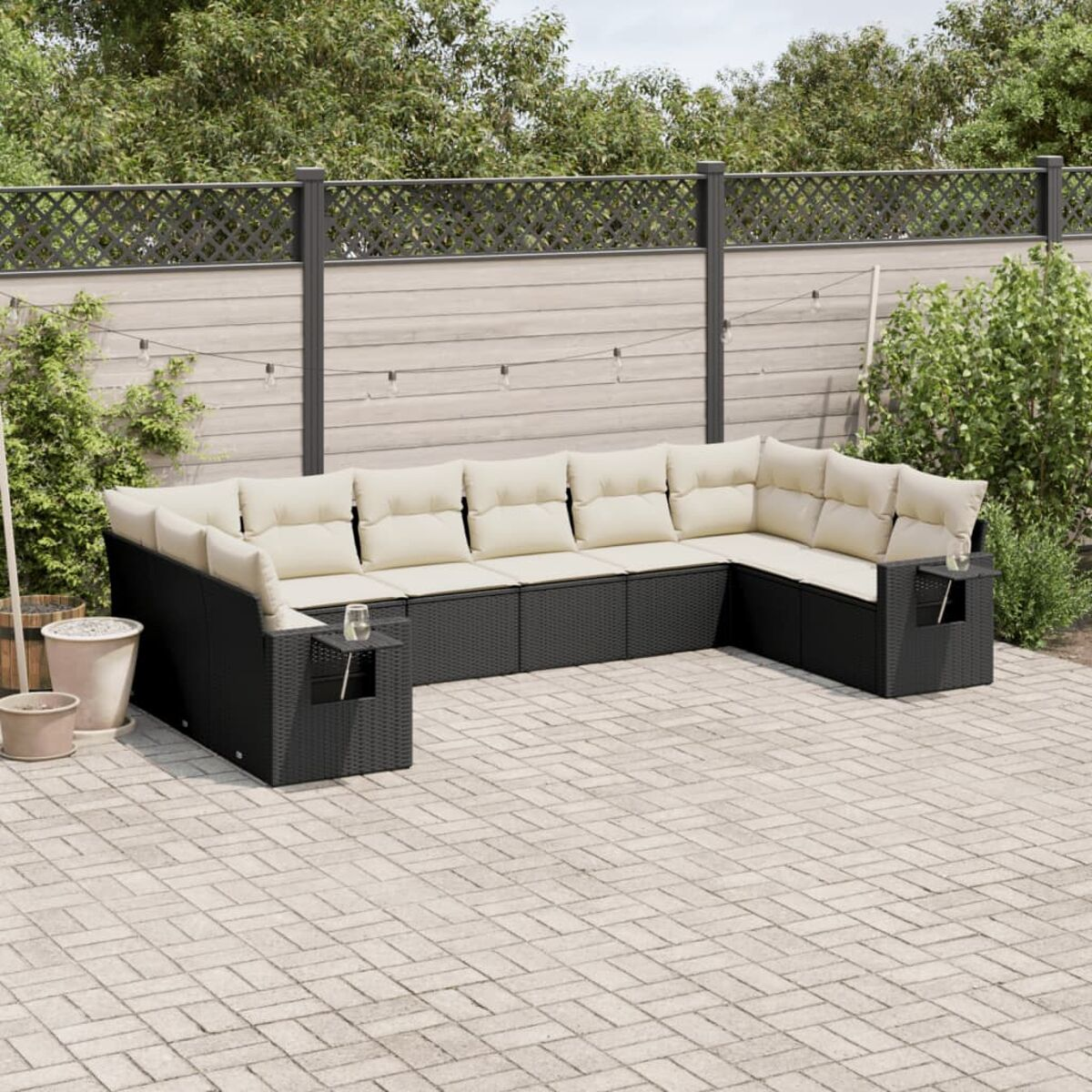 Schwarzes Rattan-Outdoor-Ecksofa mit cremefarbenen Kissen. Auf einer Backstein-Terrasse. Hintergrund enthält Zaun und Pflanzen.