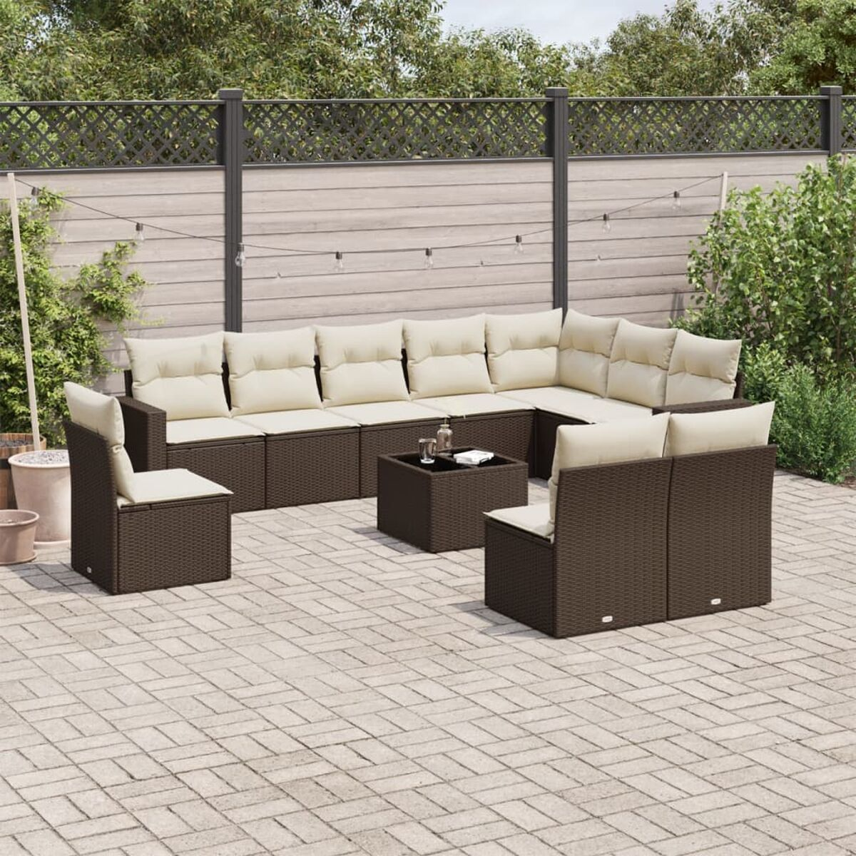 Divano angolare in vimini per esterni con cuscini color crema, tavolo e sedia su un patio con recinzione e piante.