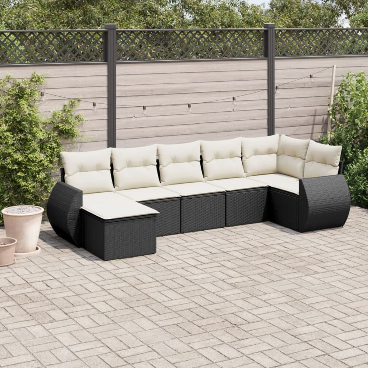 Schwarzes Wicker-Ecksofa mit cremefarbenen Kissen auf einer Terrasse. Licht und Pflanzen sind sichtbar.