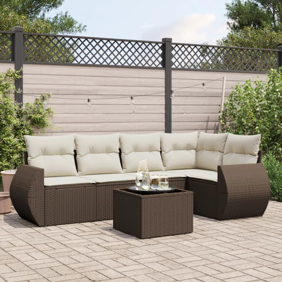 Divano componibile in vimini marrone con tavolino quadrato. Cuscini beige. Ambiente esterno con piante e luci.