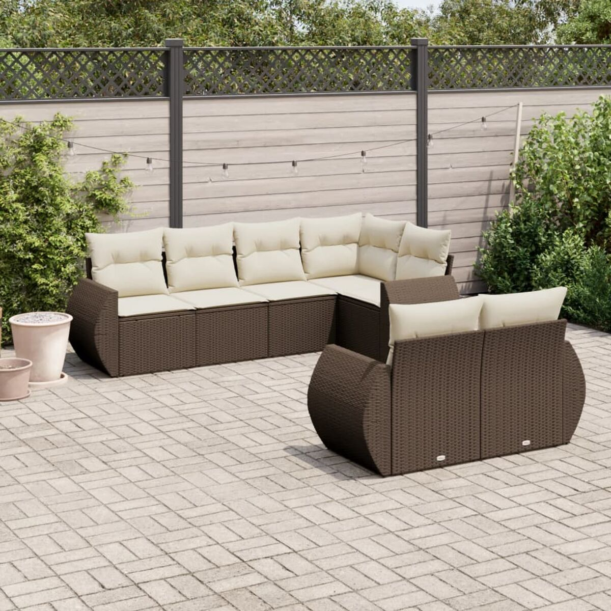 Divano angolare da esterno in vimini marrone con cuscini color crema, su un patio pavimentato. Sfondo recintato e verde.