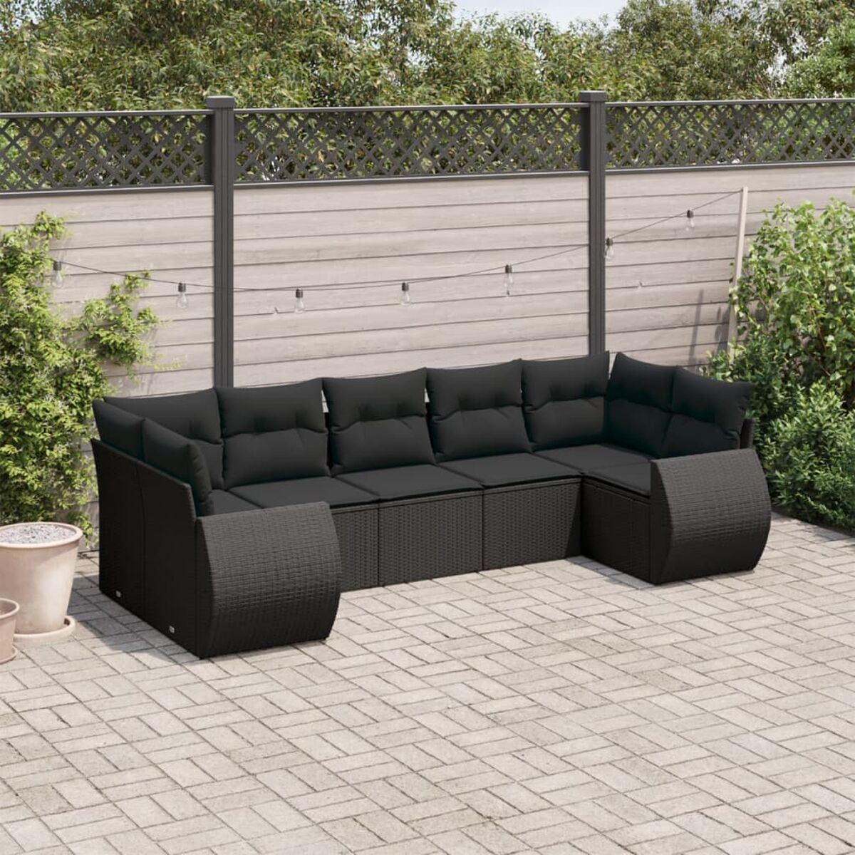 Schwarzes Ecksofa aus Korbgeflecht mit schwarzen Kissen, auf einer Ziegelterrasse angeordnet, mit Grün im Hintergrund.