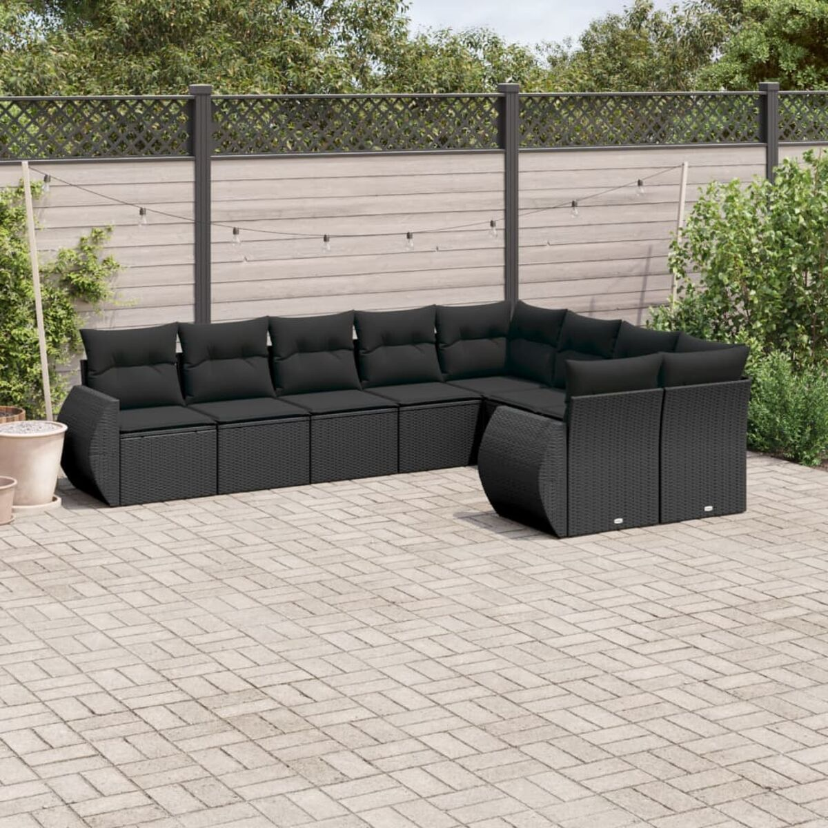 Schwarzes Ecksofa aus Korbgeflecht mit schwarzen Kissen auf einer Ziegelterrasse. Dahinter ein Zaun und Grün.