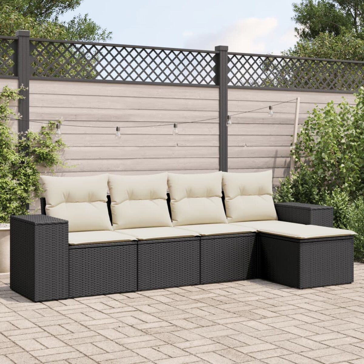 Schwarzes Rattan-Sofa mit cremefarbenen Kissen. Es steht auf einer Backstein-Terrasse. Im Hintergrund ein Zaun und Pflanzen.