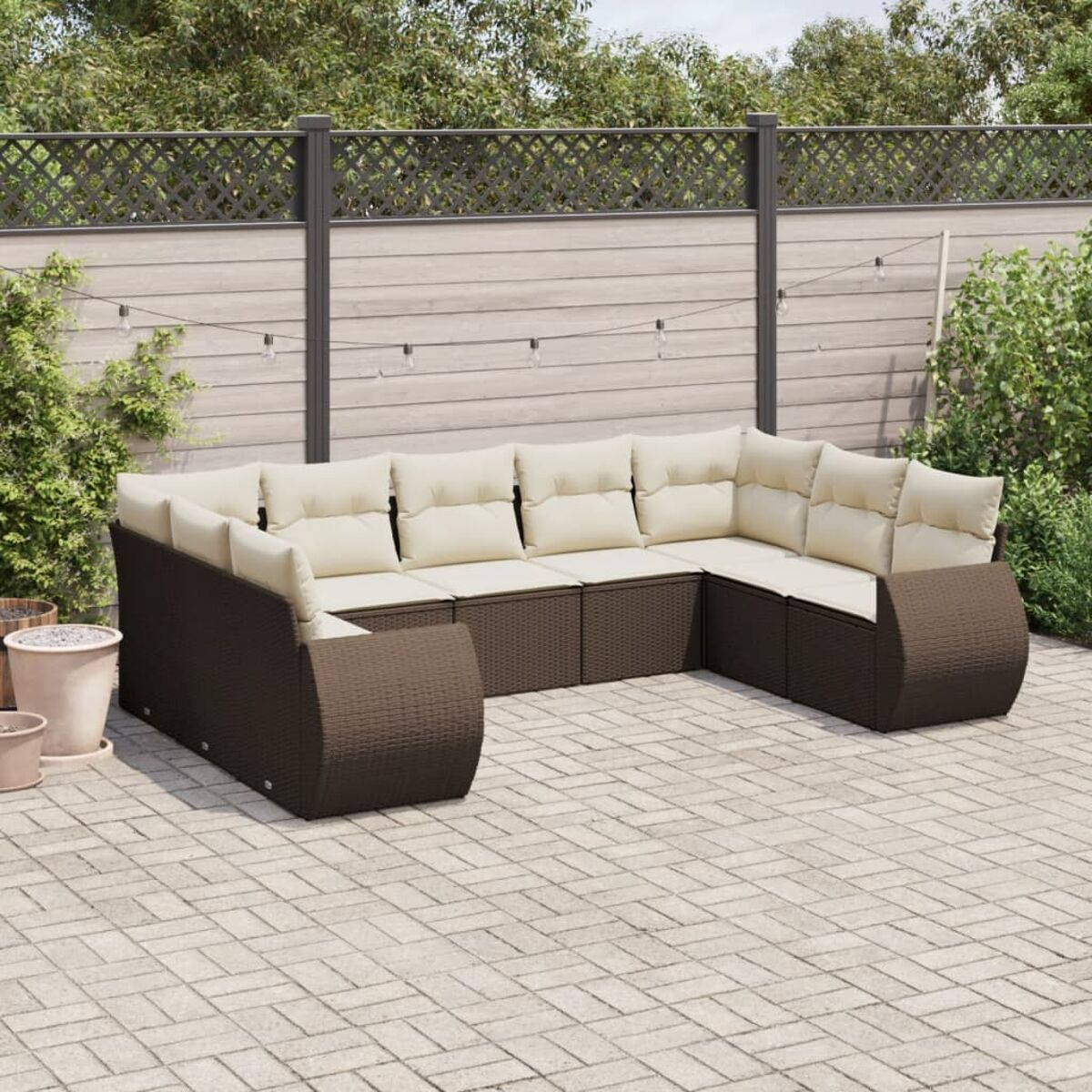 Braunes Rattan-Outdoor-Sofaset mit cremefarbenen Kissen auf Ziegelterrasse. Zaun, Pflanzen im Hintergrund.