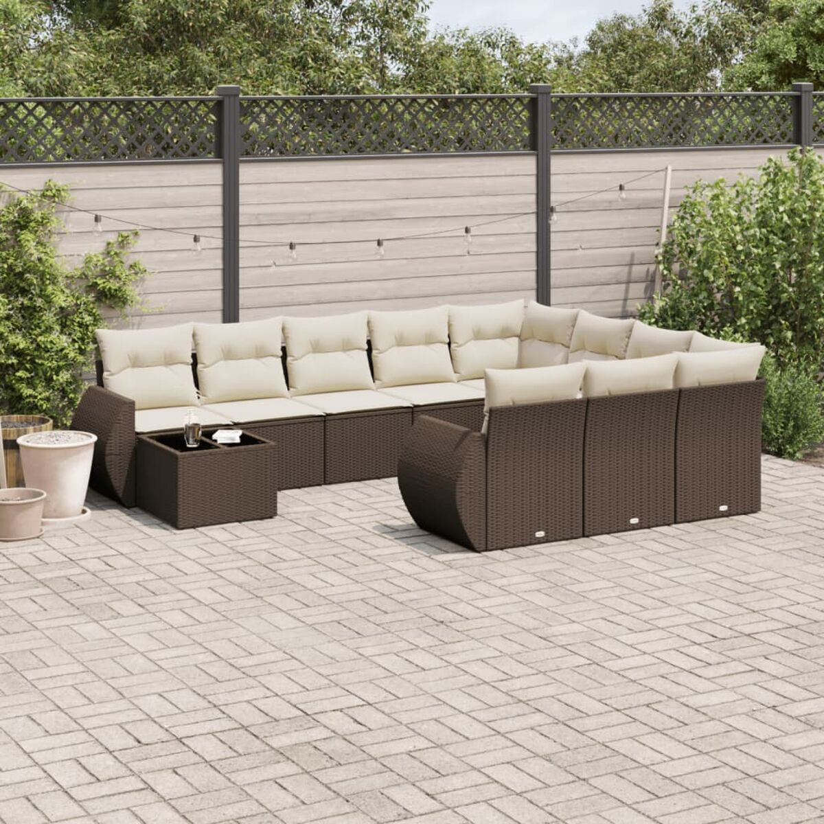Braunes Korbmöbel, cremefarbene Kissen und Tisch auf einer Ziegelterrasse. Grün und Zaun.