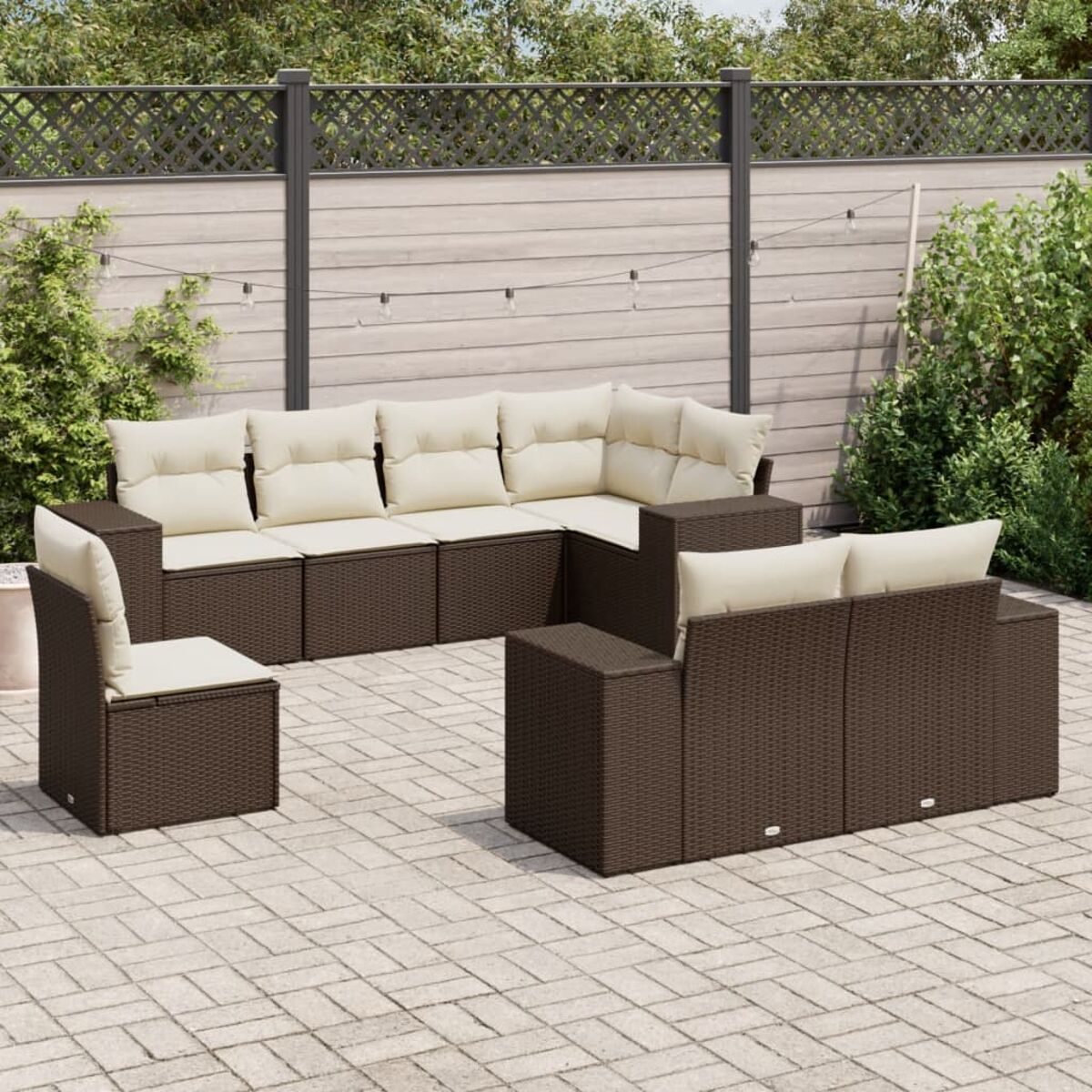 Divano angolare in rattan marrone scuro con cuscini color crema, una sedia e un tavolo su un patio in pietra. Recinzione e verde sullo sfondo.