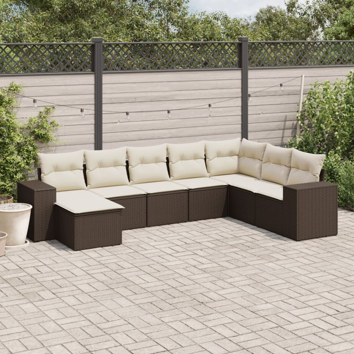 Braunes Rattan-Ecksofa mit cremefarbenen Kissen. Terrasse mit Zaun und Pflanzen im Hintergrund.