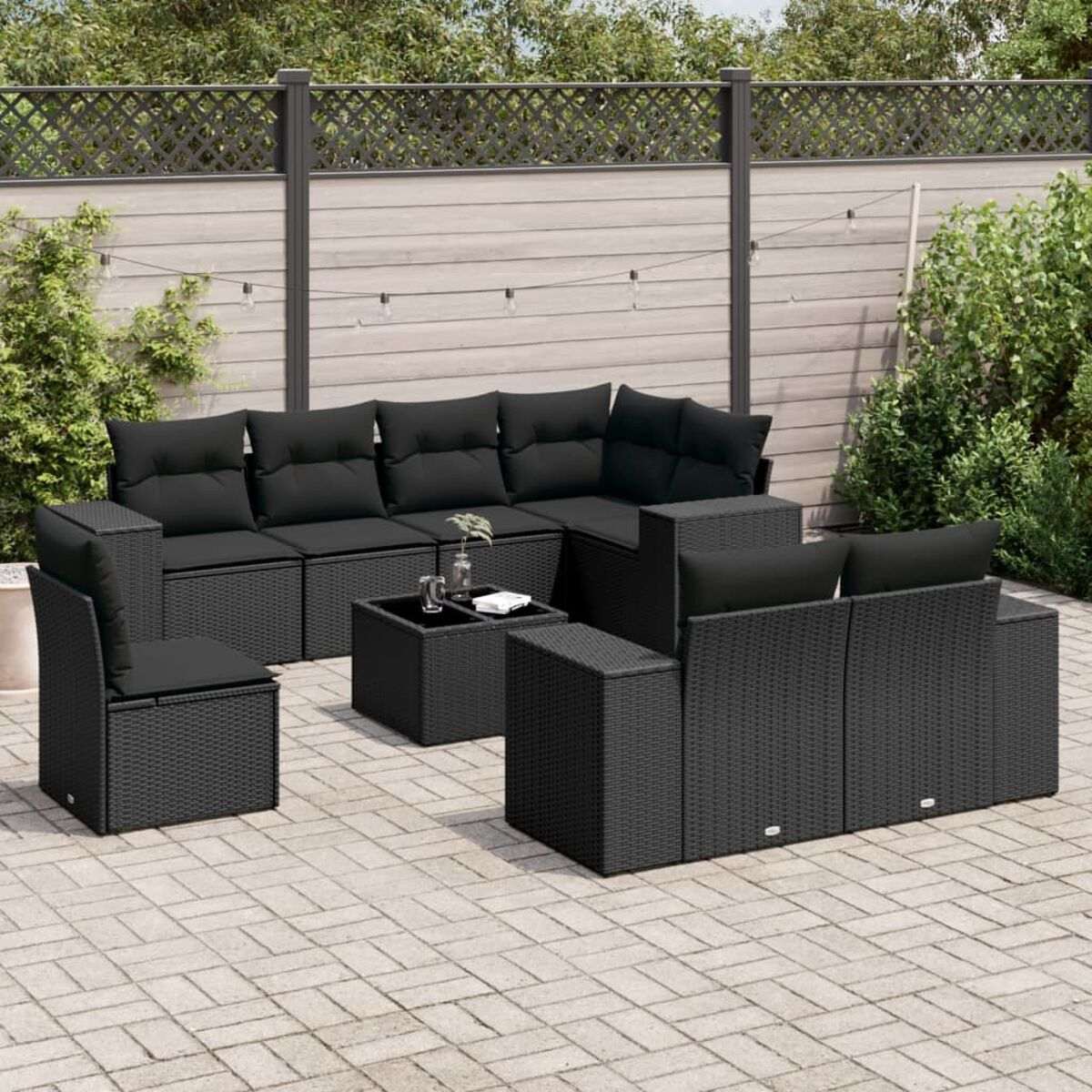 Schwarzes Weidengeflecht-Terrassenmöbel-Set. Sofa, Stühle und Tisch. Schwarze Kissen. Ziegel-Terrasse, grüner Hintergrund.