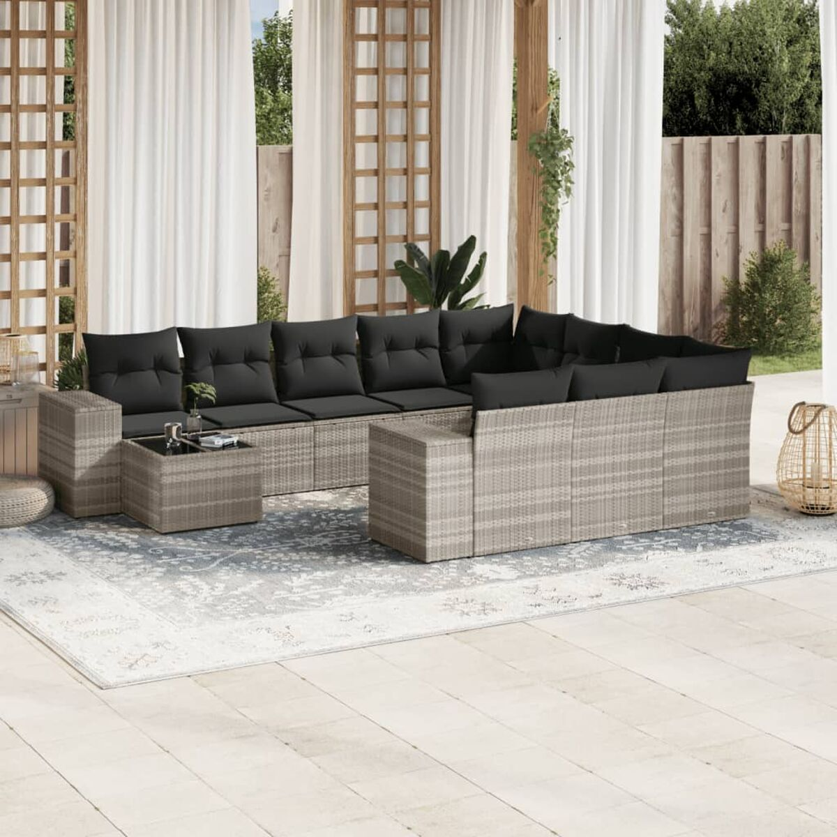 Outdoor-Ecksofa mit schwarzen Kissen, einem Tisch und einem Teppich auf der Terrasse. Weiße Vorhänge, Spalier und Pflanzen.