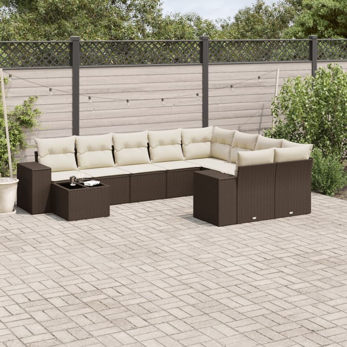 Divano angolare esterno in vimini marrone scuro con cuscini color crema. Include tavolino, su patio in mattoni e sfondo recintato.