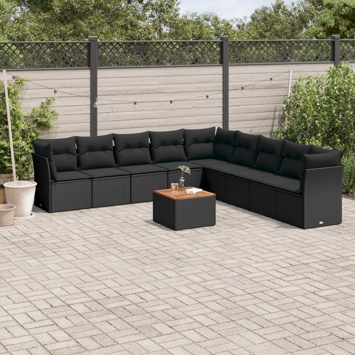 Schwarzes Korbsessel-Sofa mit schwarzen Kissen und einem kleinen Tisch auf einer Backstein-Terrasse, Zaun und Grün.