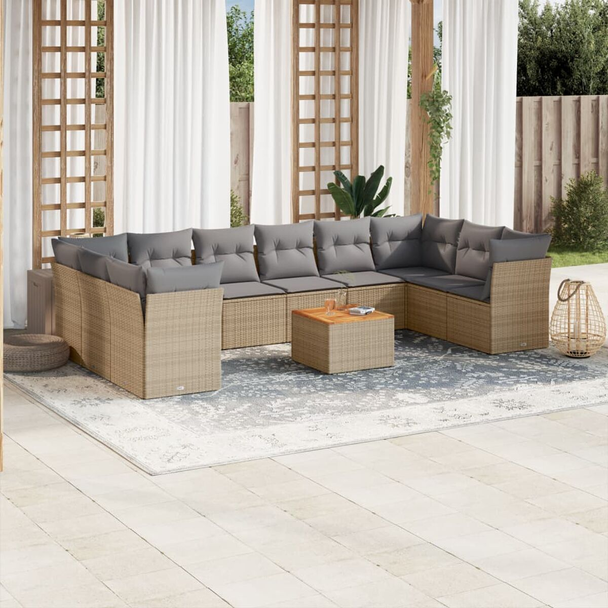 Beige Korbsitzgruppe mit grauen Kissen, Couchtisch und einem Teppich auf einer Terrasse. Vorhänge und Laub im Hintergrund.