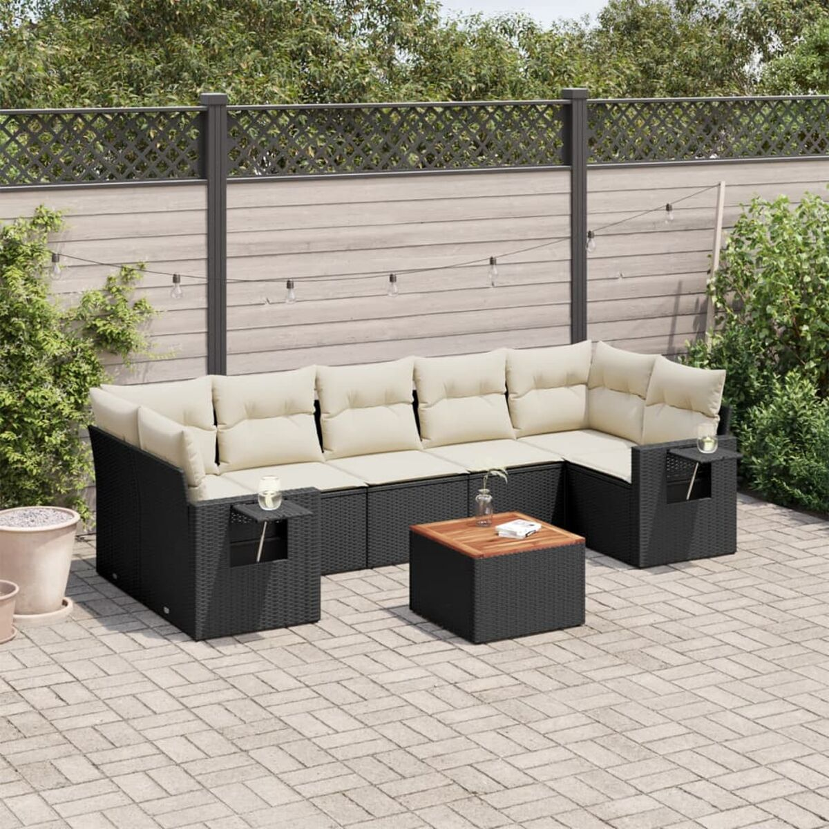 Schwarzes Ecksofa aus Korbgeflecht mit beigen Kissen und Holztisch auf Terrasse. Hintergrund Zaun.