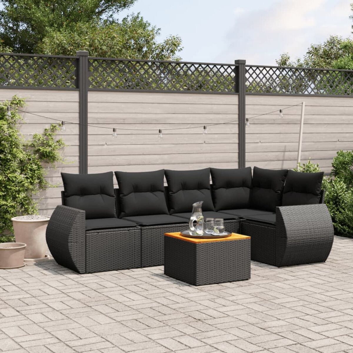 Schwarze Korbmöbel mit schwarzen Kissen, Holztisch, auf einer Steinterrasse. Laub und Zaun im Hintergrund.