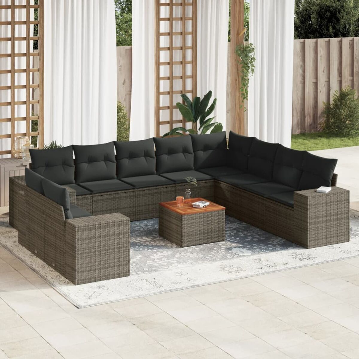 Outdoor-Rattansofa-Set mit dunklen Kissen, Couchtisch und Teppich. Weiße Vorhänge im Hintergrund.
