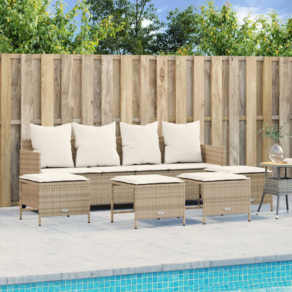 Rattan-Sofa-Set mit weißen Kissen und Tischen neben einem Pool. Zaun und Bäume im Hintergrund.