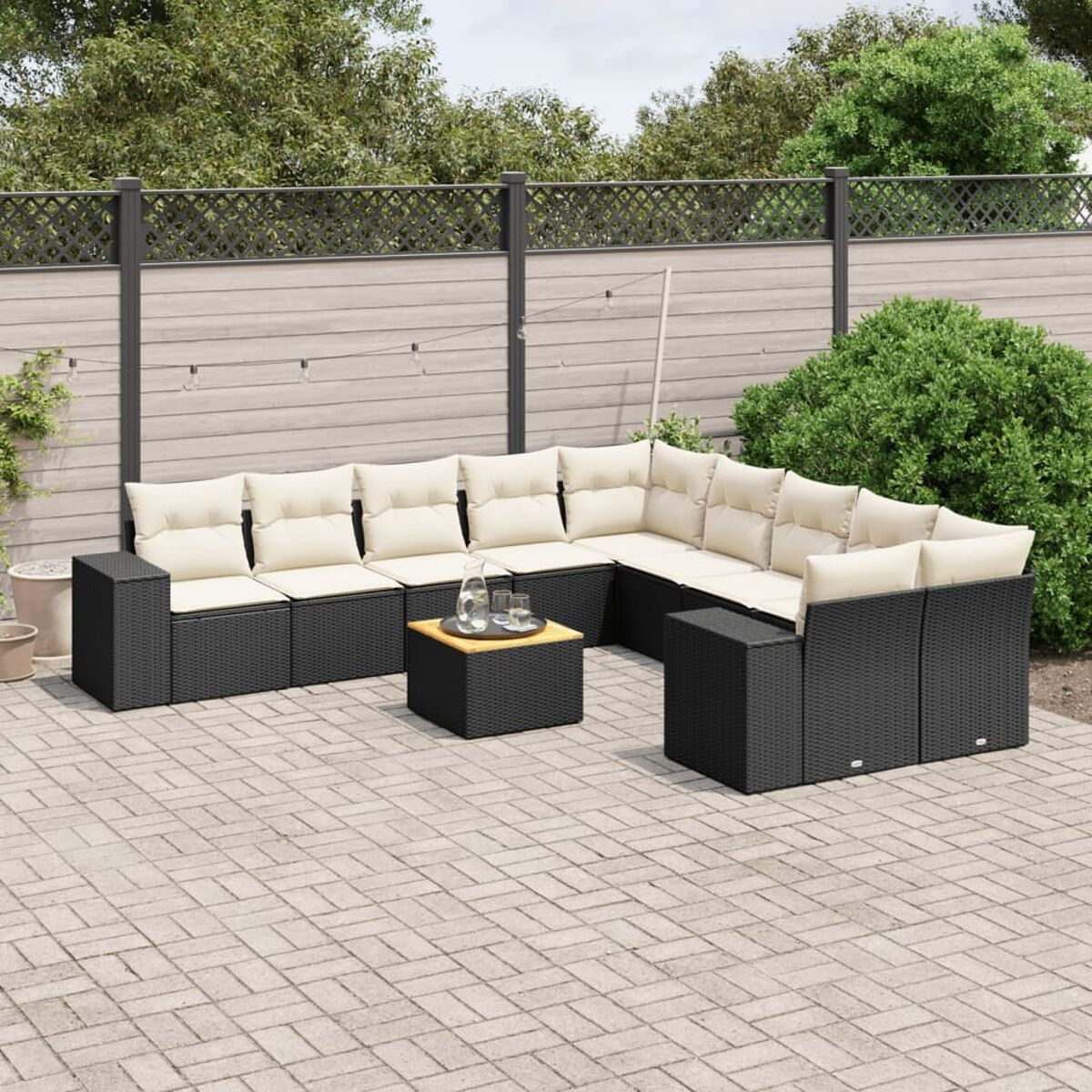 Schwarzes Ecksofa aus Korbgeflecht mit cremefarbenen Kissen und einem Tisch. Gepflasterte Terrasse mit Zaun im Hintergrund.