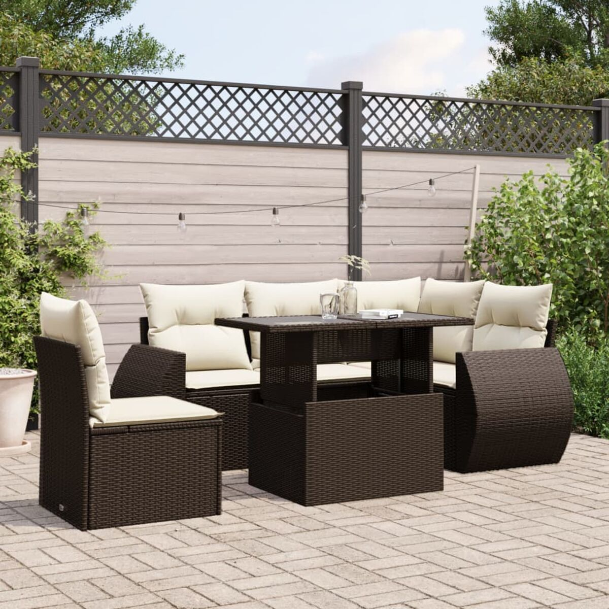 Braunes Rattan-Terrassenset mit beigen Kissen. Beinhaltet Sofa, zwei Stühle und einen Tisch. Hintergrund: Zaun und Pflanzen.