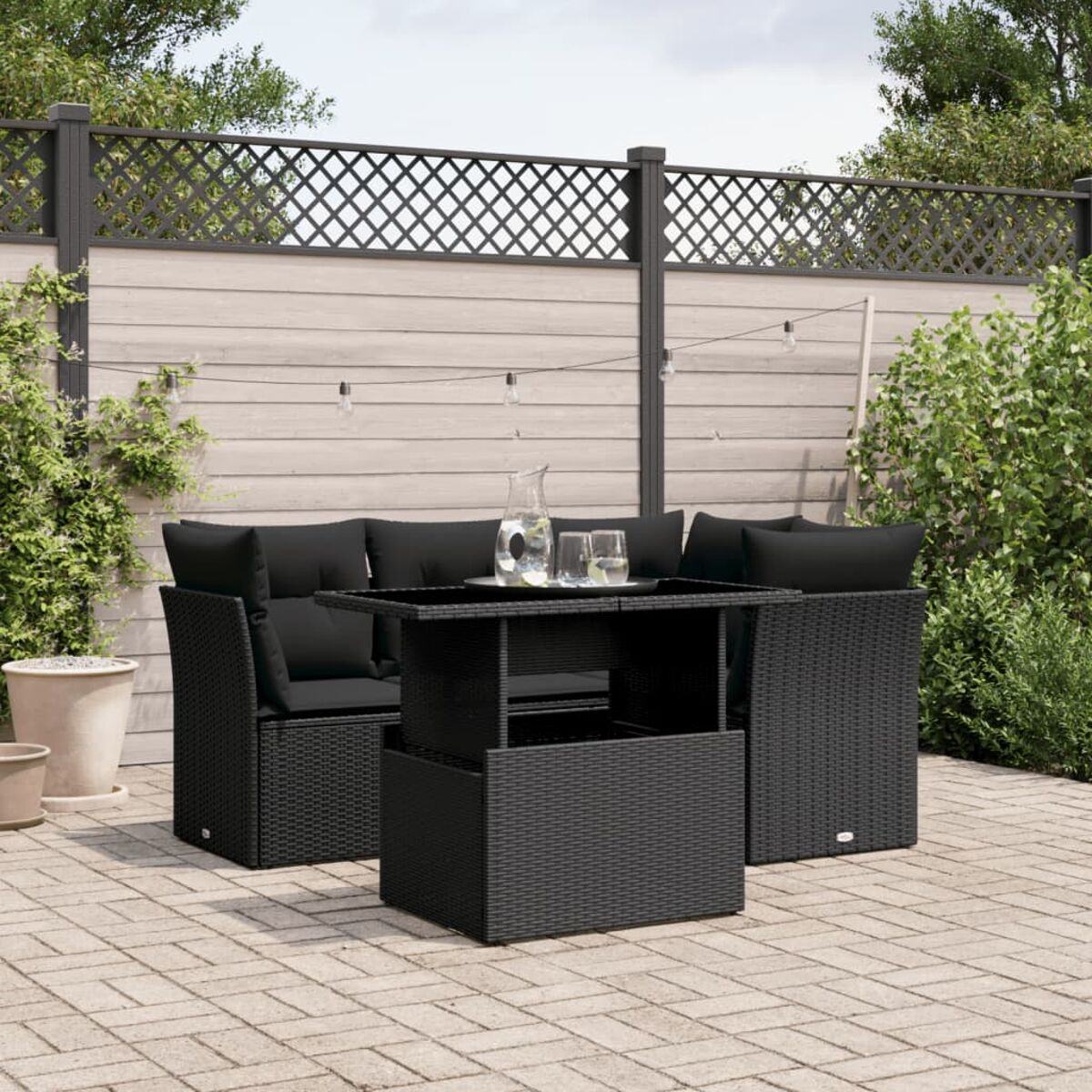 Schwarzes Gartenmöbel-Set auf einer Terrasse. Enthält Sofa, zwei Stühle und einen Tisch. Hintergrund: Zaun und Pflanzen.
