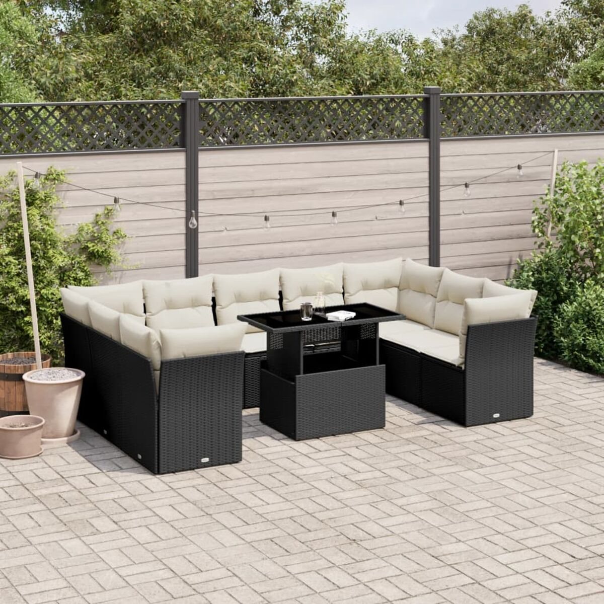 Schwarzes Rattan-Terrassenset mit beigen Kissen. Beinhaltet ein Ecksofa und einen Tisch. Hintergrund: Zaun.