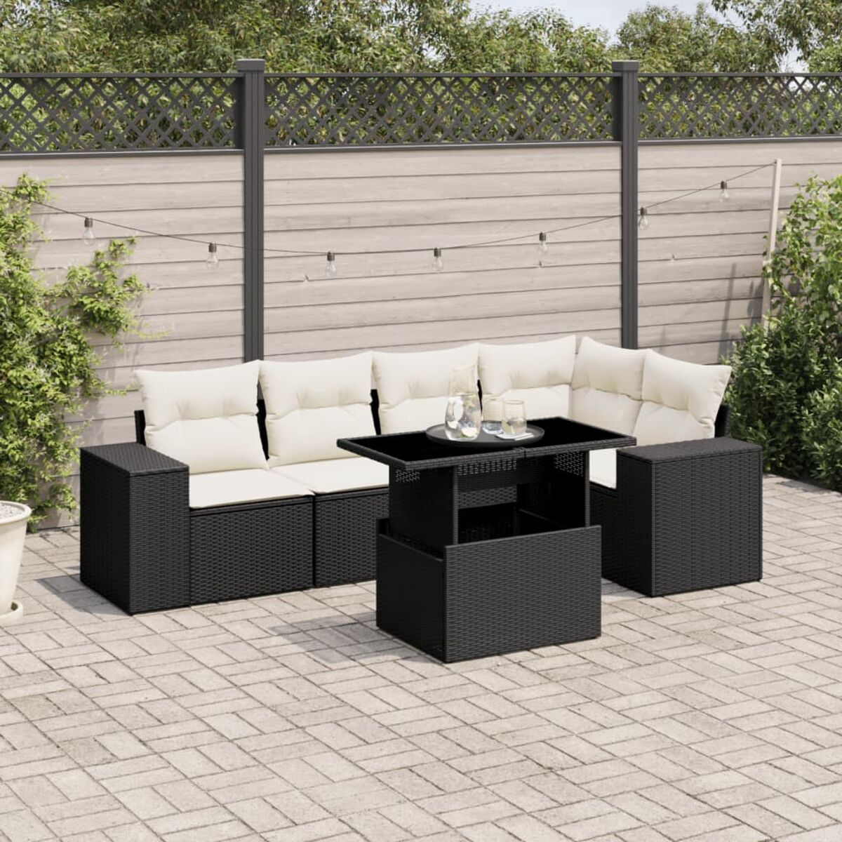 Schwarzes Outdoor-Sofa aus Korbgeflecht mit weißen Kissen und einem schwarzen Tisch. Ein grauer Zaun ist im Hintergrund.