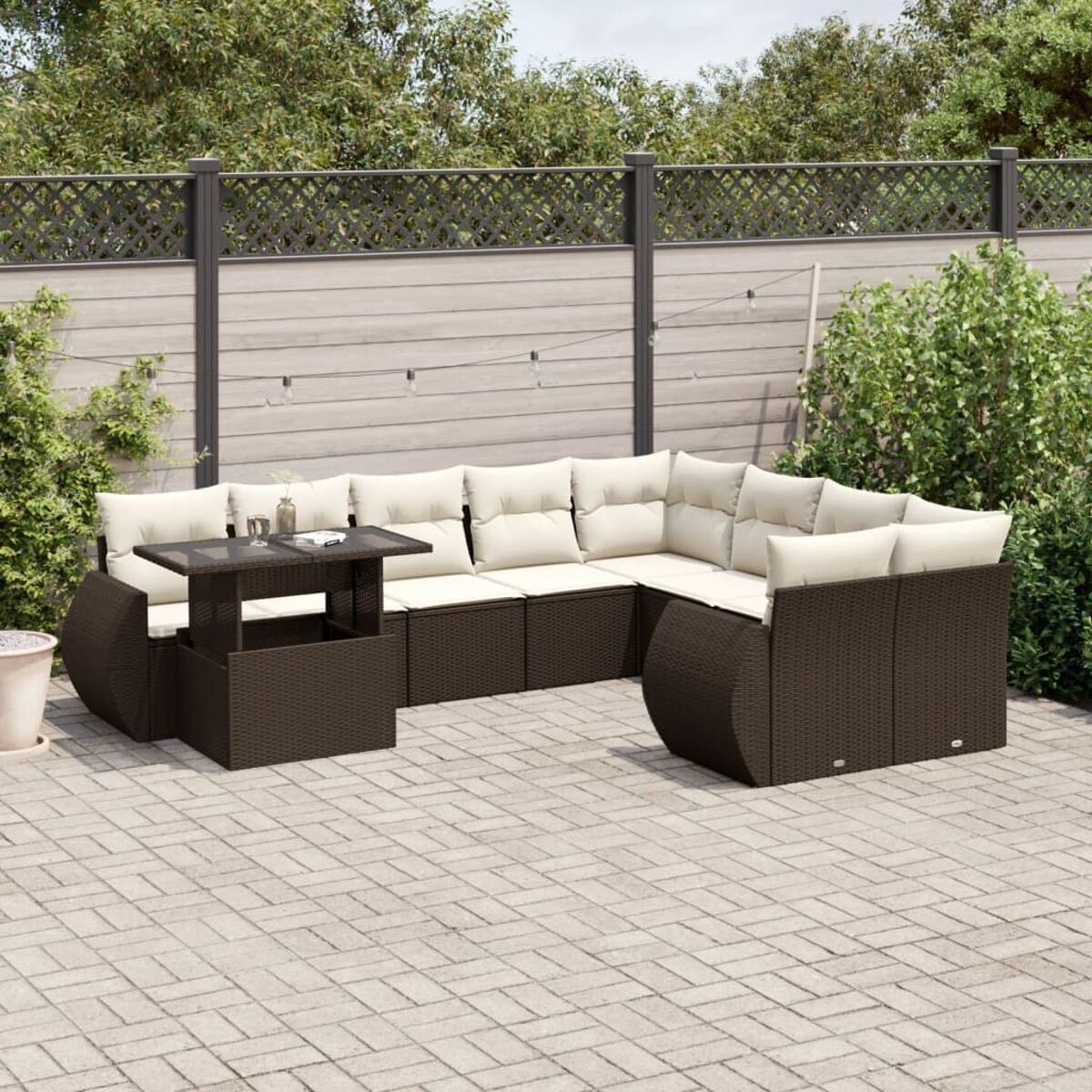 Divano componibile da esterno in rattan marrone con cuscini beige e un tavolo. Patio in mattoni con verde sullo sfondo.