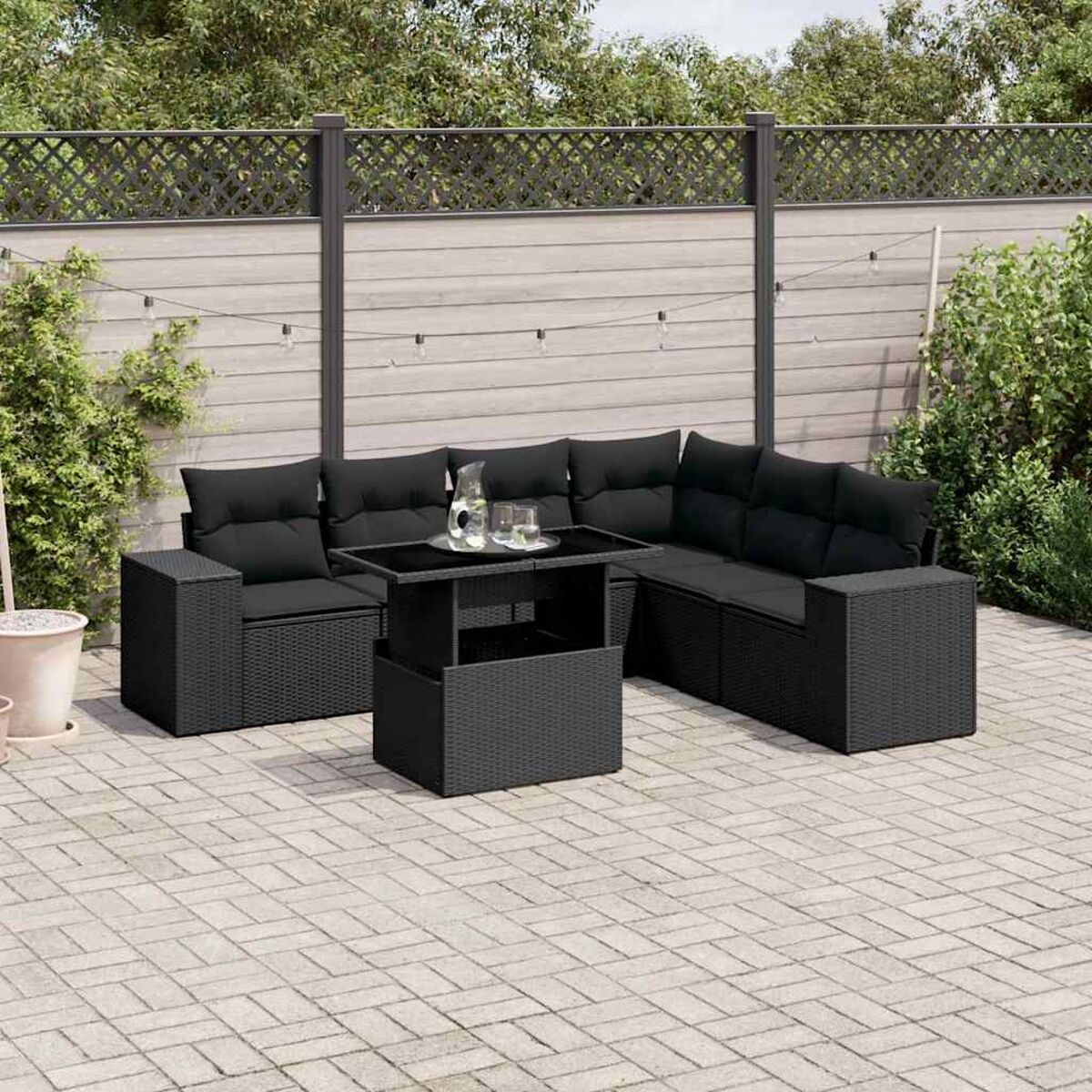 Divano componibile in vimini nero con tavolo in vetro. Patio con pavimento in mattoni, recinzione e piante.