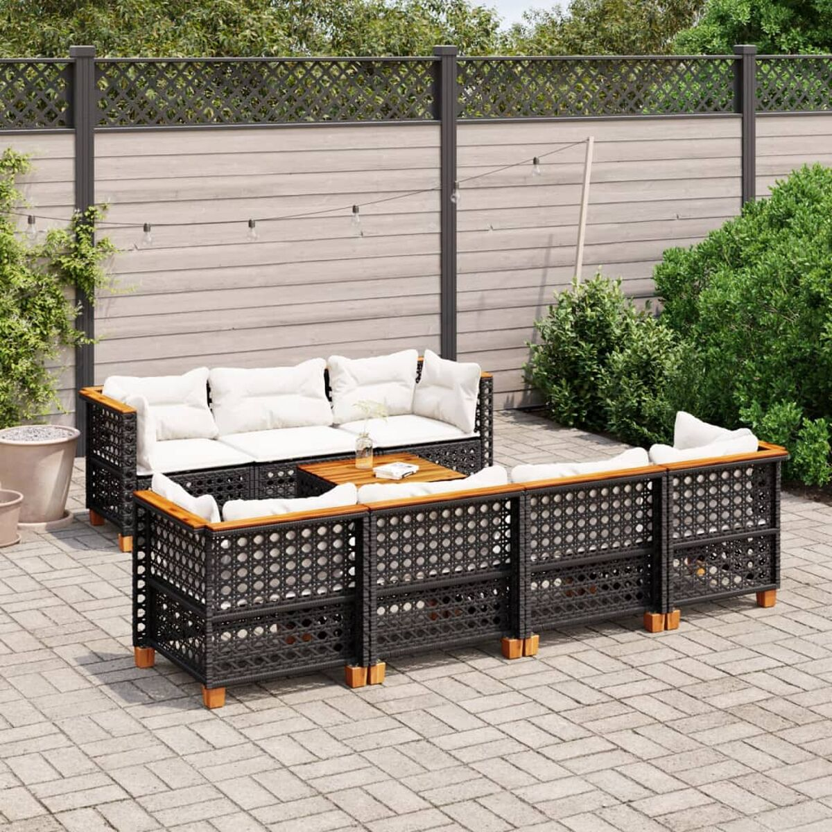 Gartenmöbelset, schwarzer gewebter Rahmen, weiße Kissen. Holztisch. Garten mit Zaun und Grün.
