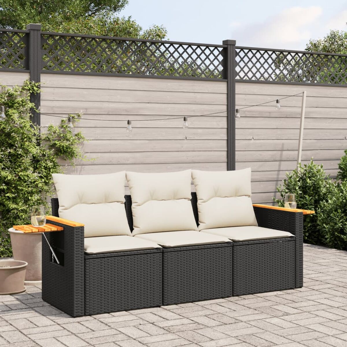 Schwarzes Rattan-Sofa mit cremefarbenen Kissen, Holzarmlehnen und Getränken. Hintergrund: Zaun und Pflanzen.